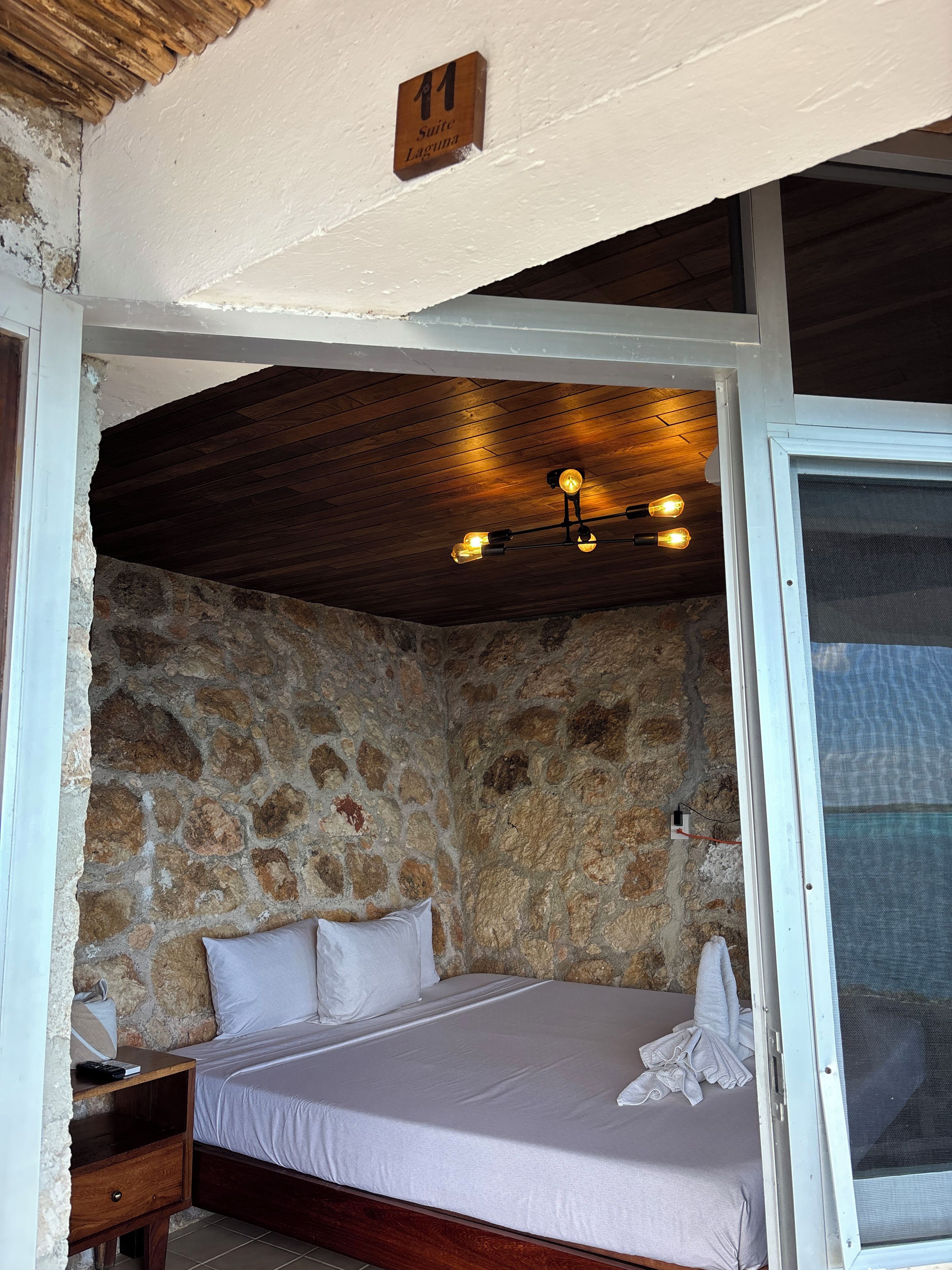 Una cama individual en una habitación con paredes de piedra, techo de madera y una mesita de noche pequeña.