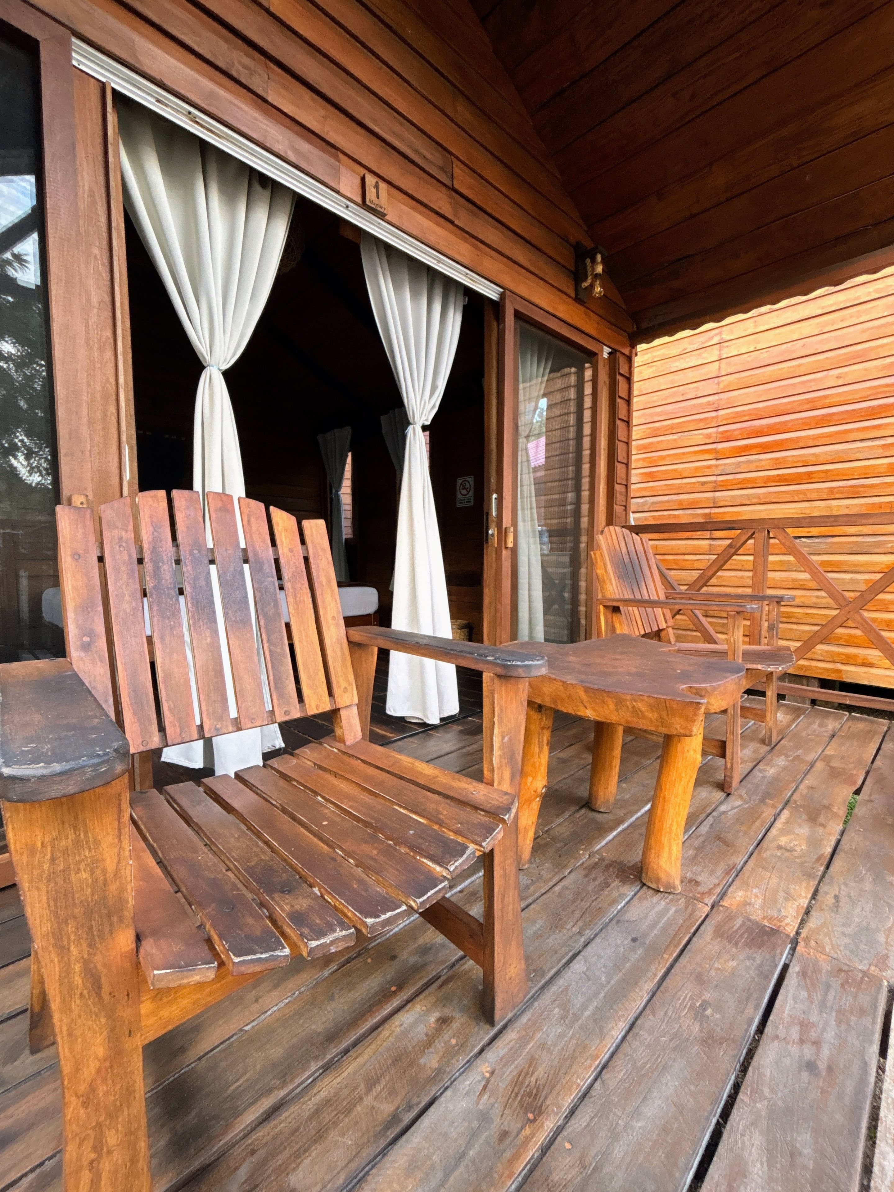 Una terraza de madera con una mesa y dos sillas, un ventanal corredero con cortinas y un muro de madera.