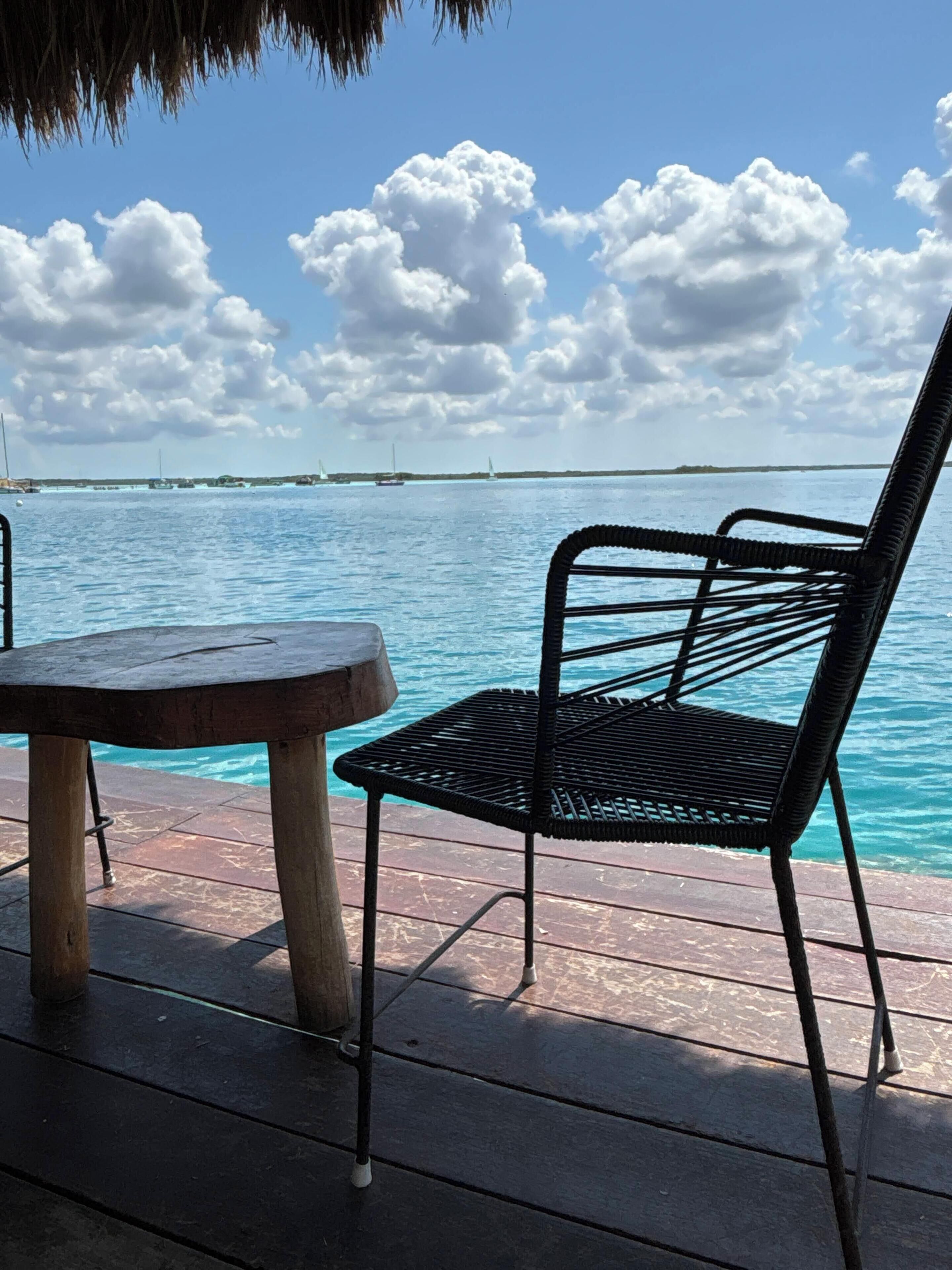 A wooden table and a metal chair on a wooden deck overlooking a body of water with sailboats.