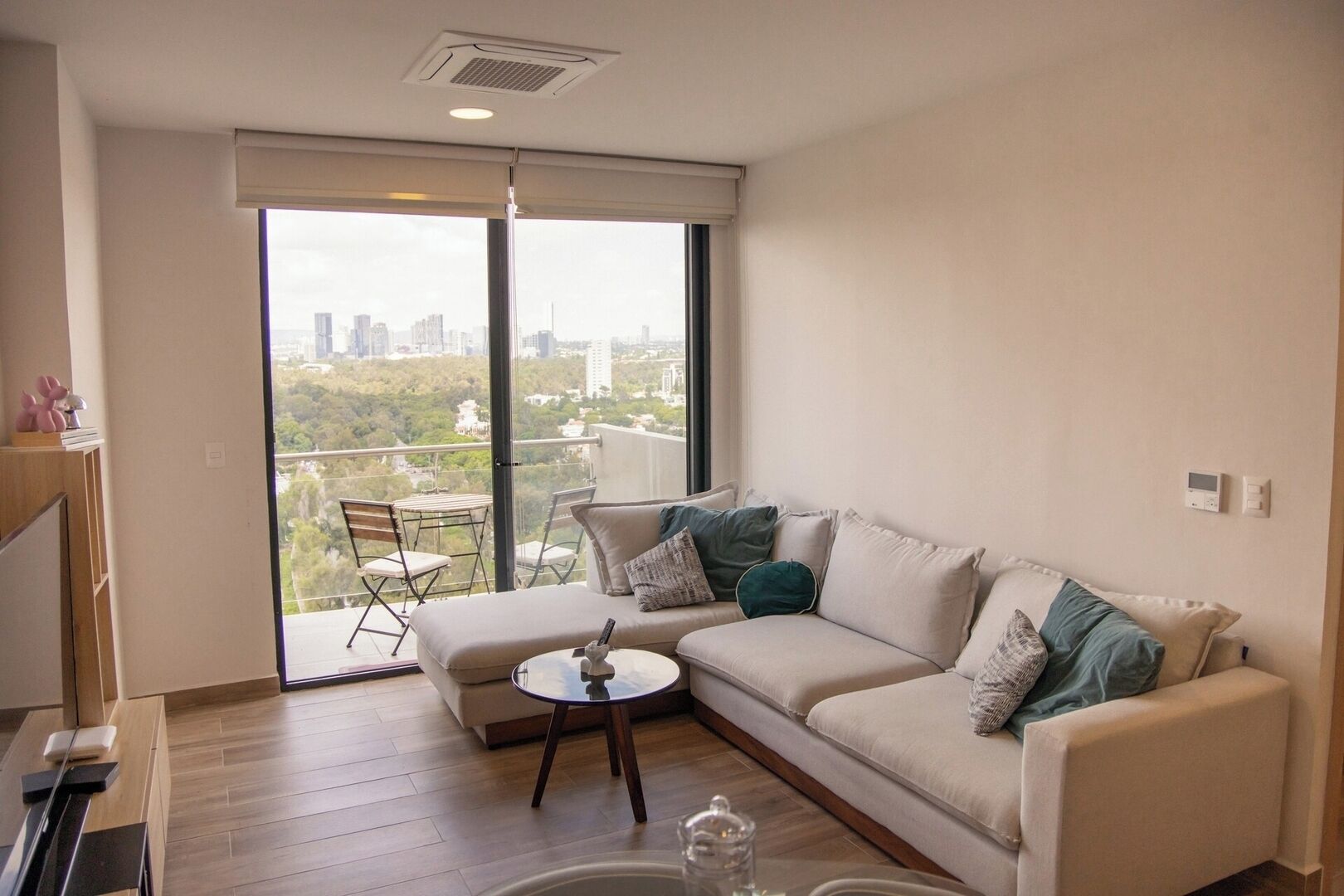 Ein modernes Wohnzimmer mit einem beigefarbenen Sofa, einem Glastisch und Blick auf eine Stadtlandschaft durch große Fenster.