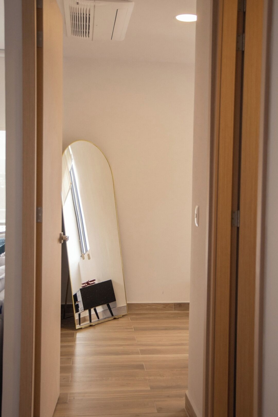 A hallway with a wooden floor, a door on the right, and a large mirror on the left.