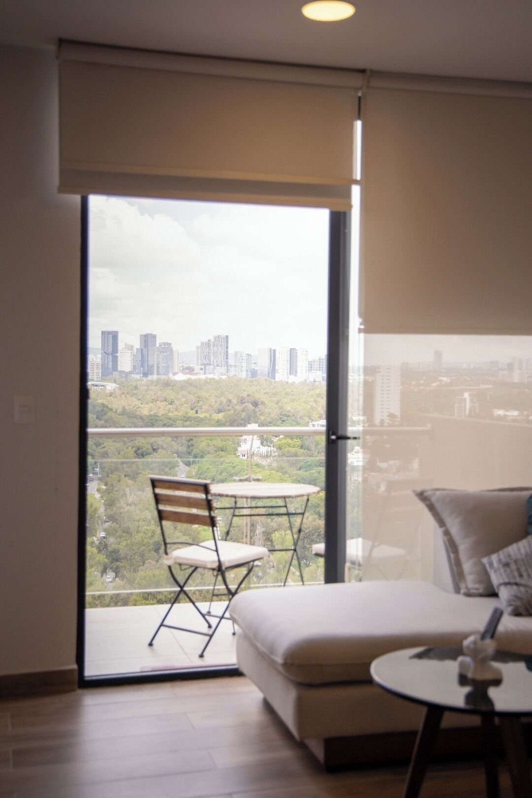 A modern living room with a sofa, a small round table, and a chair. A sliding glass door leads to a balcony with a city view.