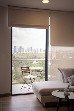 A modern living room with a sofa, a small round table, and a chair. A sliding glass door leads to a balcony with a city view. - Elegant 2-Bedroom Condo in Colomos, Providencia (Guadalajara)
