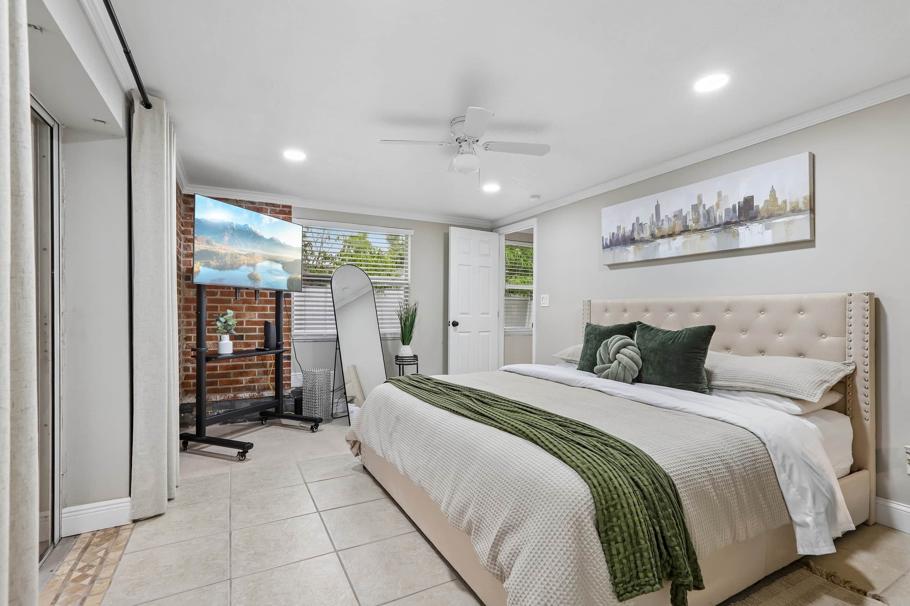 A bedroom with a bed, a cityscape painting, a ceiling fan, and a brick wall.