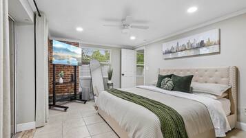 A bedroom with a bed, a cityscape painting, a ceiling fan, and a brick wall.