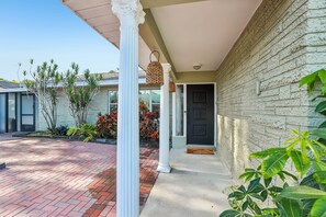 A covered porch with a brick pathway leading to a door, surrounded by greenery and a brick wall. - Resort Pool Home • 3 Min to Beach • Pool Table (Pinellas County)