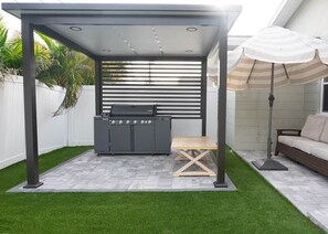 A covered patio area with a grill, a wooden table, and a striped umbrella. - Pool, Mini Golf, Pool Table, Fire Pit, Sleeps 10 (Pinellas County)