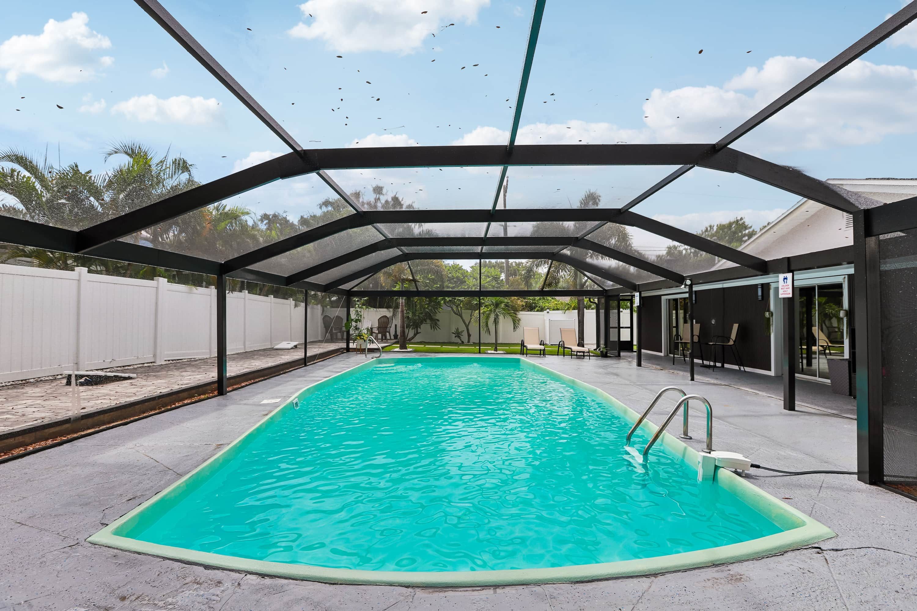 A covered swimming pool with a clear blue water surface, surrounded by a white fence and a covered patio area.