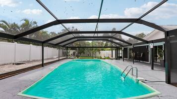A covered swimming pool with a clear blue water surface, surrounded by a white fence and a covered patio area.