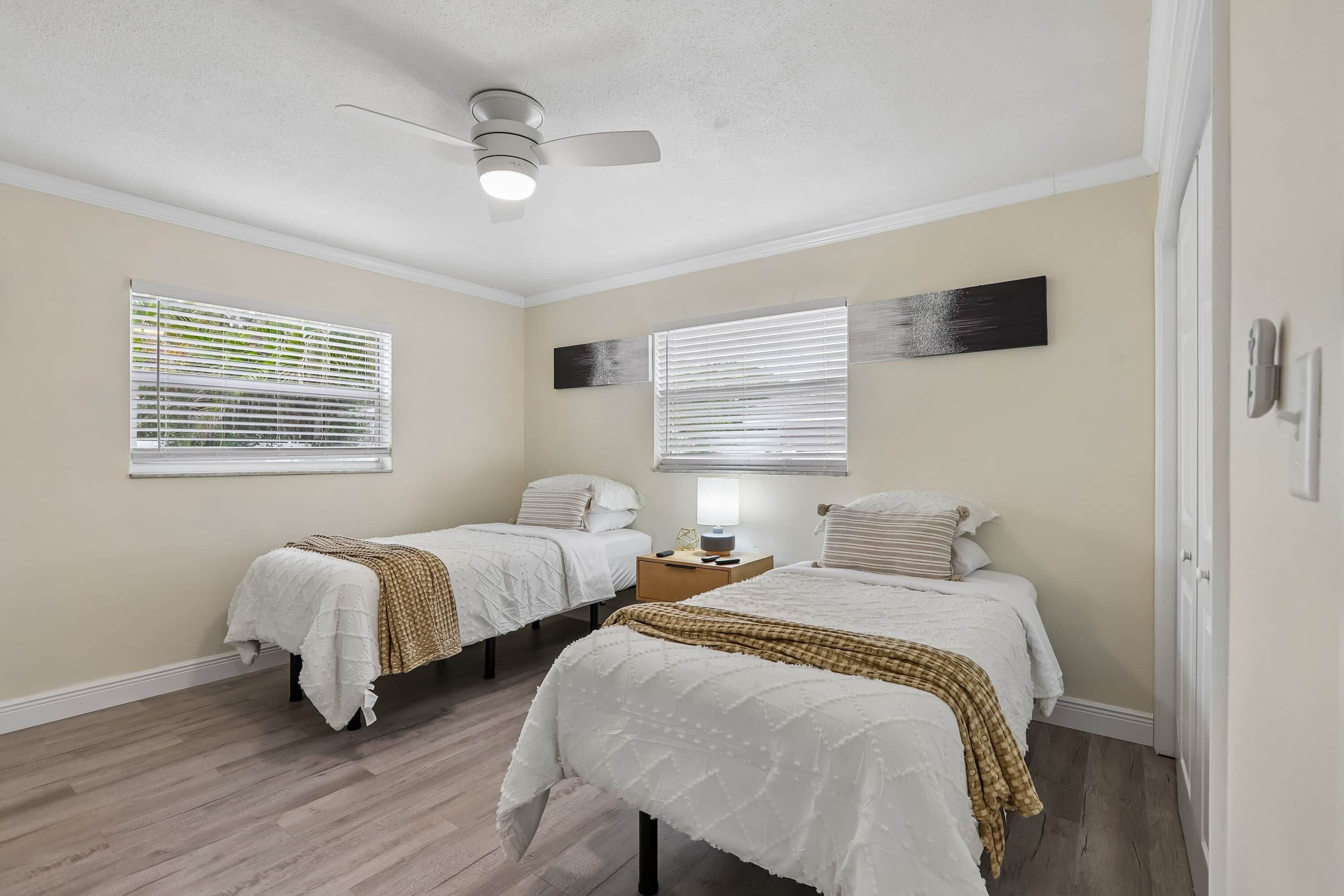 A bedroom with two beds, a ceiling fan, and two windows with blinds.