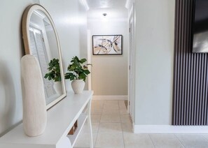 A hallway with a white console table, a large mirror, two potted plants, and a framed abstract painting on the wall. - Pool, Mini Golf, Pool Table, Fire Pit, Sleeps 10 (Pinellas County)