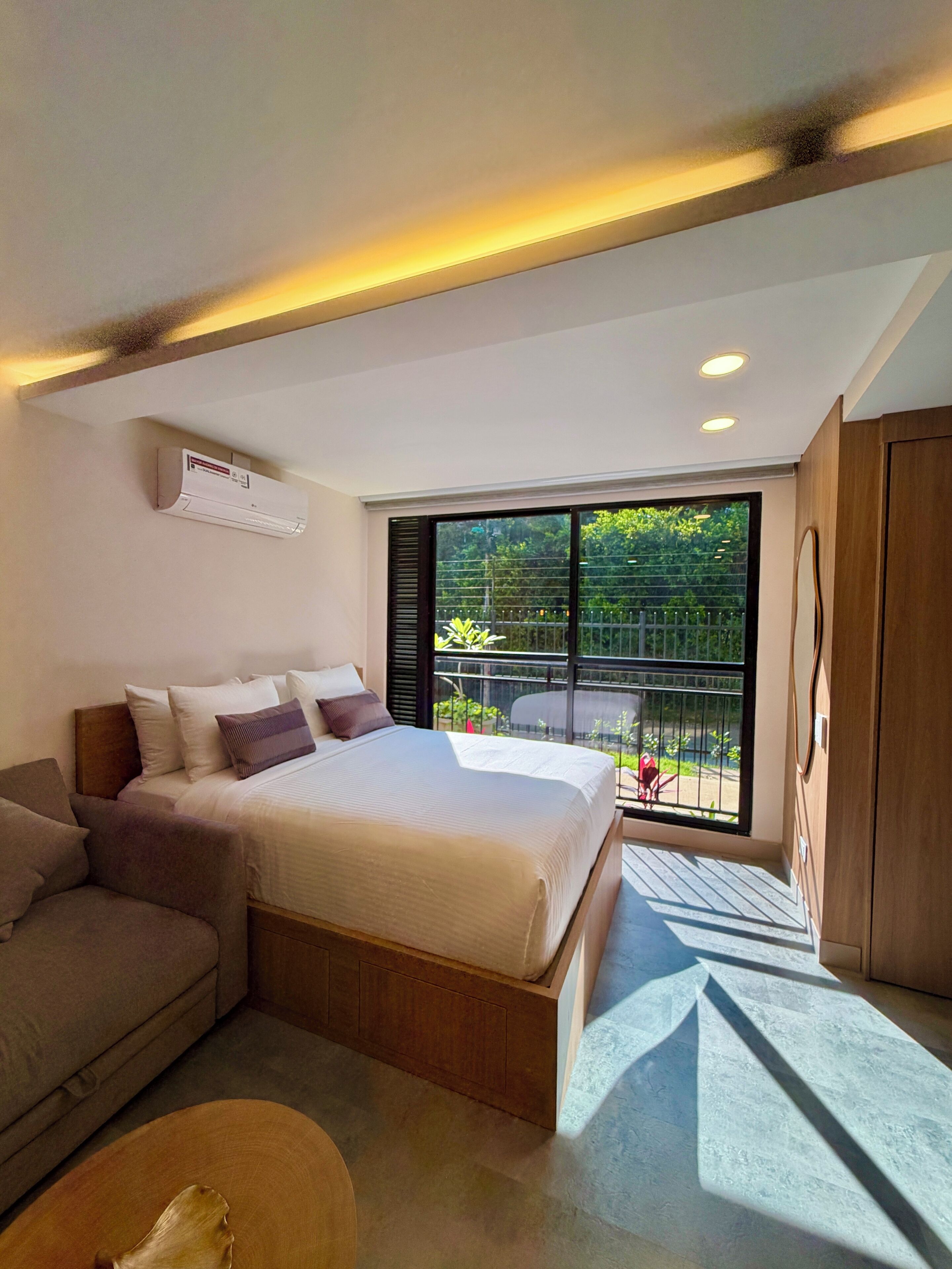 Ein modernes Schlafzimmer mit Bett, einer Couch, einem Holzschrank und einem Balkon mit Blick auf Grünflächen.