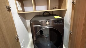 A compact laundry room with a black washing machine, wooden shelving above, and a tiled floor. - ApartaEstudio Pance - Wifi y Coworking (Cali)