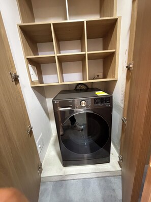 A compact laundry room with a black washing machine, wooden shelving above, and a tiled floor. - ApartaEstudio Pance - Wifi y Coworking (Cali)