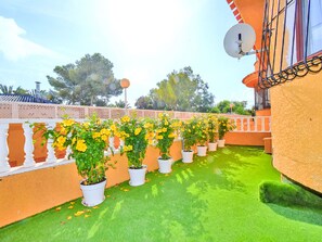 Terraço com vasos de plantas amarelas floridas dispostos em linha, uma antena parabólica e gramado sintético verde.