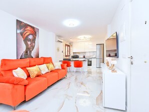 Uma sala de estar moderna com um sofá laranja vibrante, uma cozinha ao fundo e um armário branco com uma televisão de tela plana.