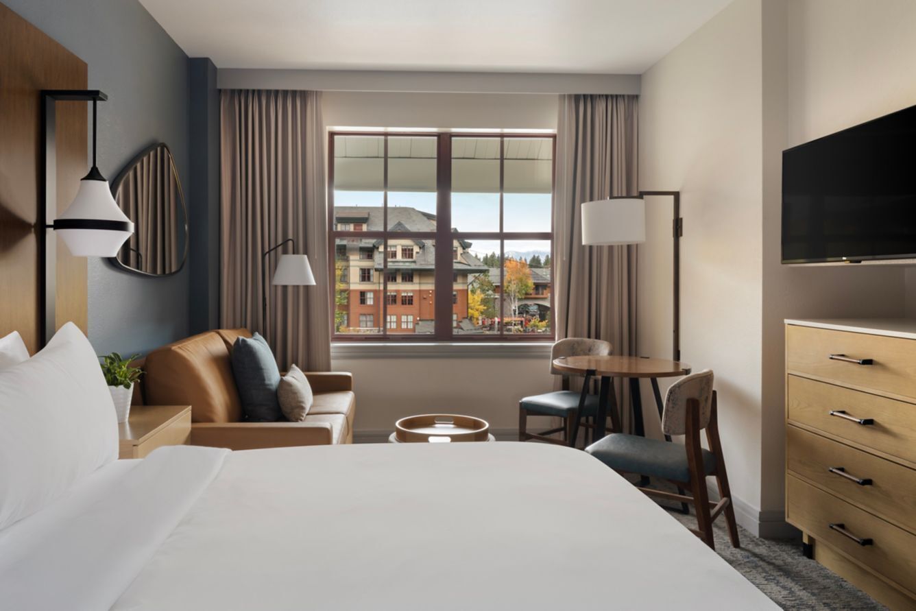 Una habitación de hotel con cama, escritorio, silla, televisión y ventana con cortinas.