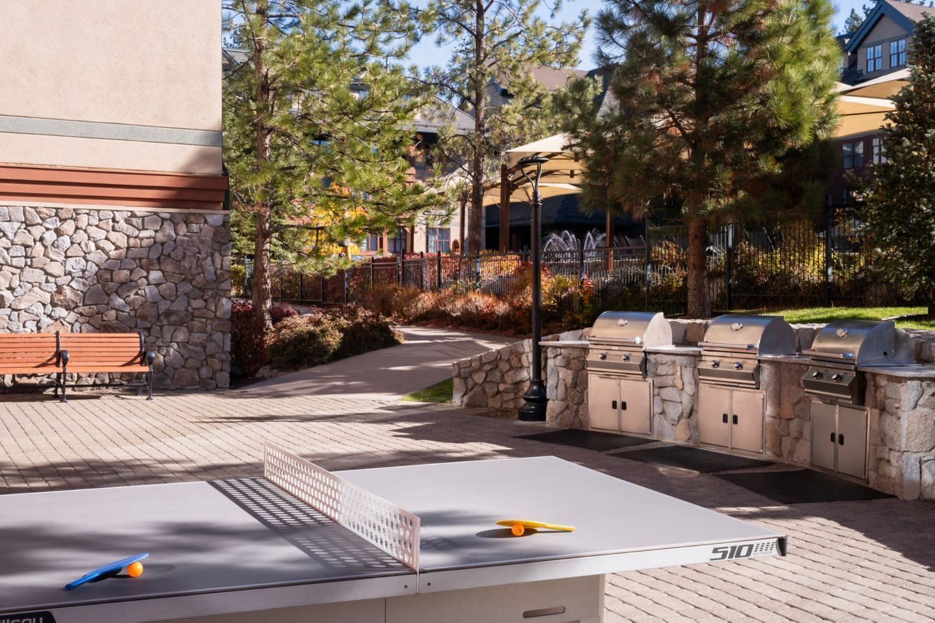 An outdoor patio with a ping pong table, a stone wall, a bench, and a barbecue grill.