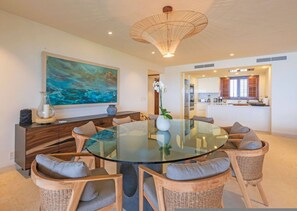 A modern dining area with a glass table surrounded by wicker chairs, a large abstract painting on the wall, and a kitchen in the background. - Condo Las Olas 4BD - Tau Punta Mita (Punta de Mita)
