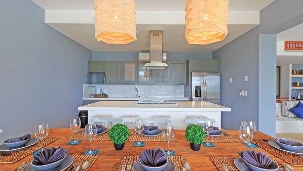 A modern kitchen with a central island, two pendant lights, and a dining area set for six with placemats and tableware. - Condo Terramar 4BD - Terrazas Punta Mita (Punta de Mita)