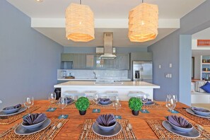 A modern kitchen with a central island, two pendant lights, and a dining area set for six with placemats and tableware. - Condo Terramar 4BD - Terrazas Punta Mita (Punta de Mita)