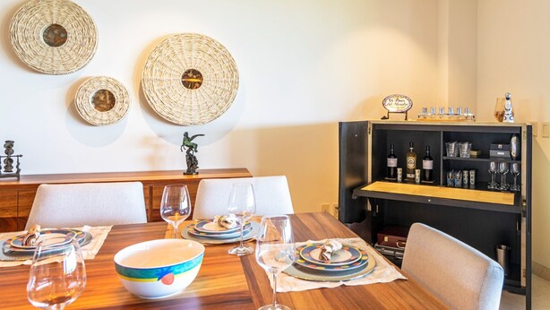 A dining area with a wooden table set for four, surrounded by chairs. Above the table, there are three woven wall decorations. To the right, there's a black cabinet with wine bottles and glasses. - Condo Balboa 3BD - Tau Punta Mita (Punta de Mita)