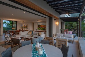 A spacious living area with a dining table, sofas, and chairs, a kitchen area with a dining table and chairs, and a large window with a view of palm trees. - Condo Piscis 3BD - Marietas Punta Mita (Punta de Mita)
