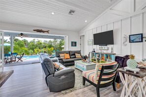 A spacious living area with a pool view, leather sofas, a flat-screen TV, and various decorative items. - Coastal Bokeelia Retreat | Zero-Entry Pool + Boat Trailer Parking Near Pineland Marina (Bokeelia)