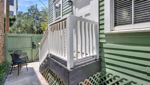 Ein Wohnhaus mit grüner Fassade, einer weißen Veranda mit Geländer und einer kleinen Außenbereich-Sitzgruppe mit Stühlen.
