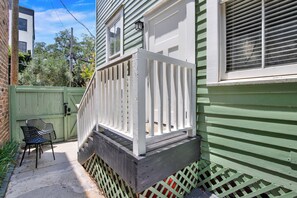 A residential property with a green exterior, a white front porch with a railing, and a small outdoor seating area with chairs. - Bright Modern Retreat in Savannah's Historic District | Heated Pool Access (Savannah)