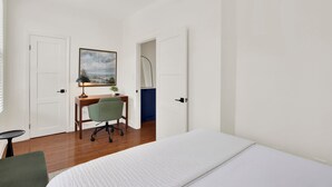 Ein Schlafzimmer mit Bett, Schreibtisch mit Stuhl, einem Bild an der Wand und einer Tür zu einem anderen Raum.