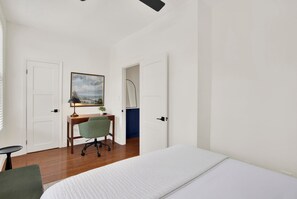 A bedroom with a bed, a desk with a chair, a painting on the wall, and a door leading to another room. - Bright Modern Retreat in Savannah's Historic District | Heated Pool Access (Savannah)