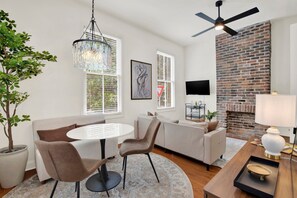 A modern living room with a round table, two chairs, a sofa, a ceiling fan, a brick wall, and a television. - Bright Modern Retreat in Savannah's Historic District | Heated Pool Access (Savannah)