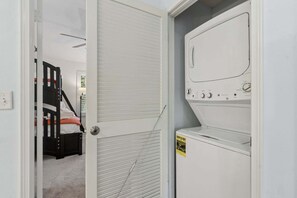Ein Waschraum mit einer Frontlader-Waschmaschine und -trockner, einer Tür mit Fenster und einem Kinderstockbett im angrenzenden Zimmer.
