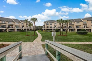 A pathway leading to a row of multi-story buildings with balconies, surrounded by well-maintained grass and palm trees. - Sailmaker Gated Complex: Near Pool & Beach (Fernandina Beach)