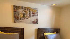 Two single beds with wooden headboards, a painting of a street scene, and a ceiling light fixture.