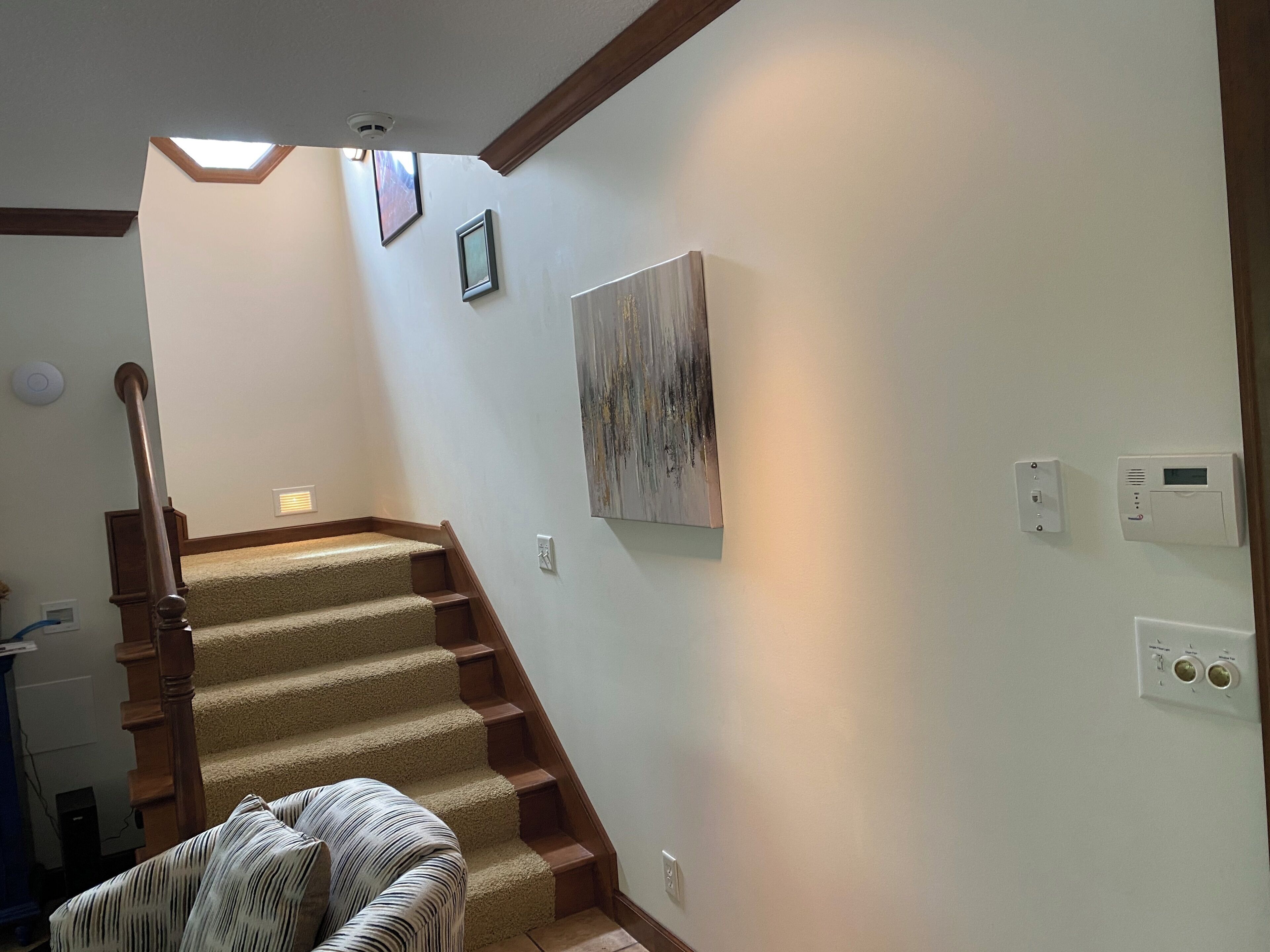 Un escalier avec des marches recouvertes de moquette et des rampes en bois, menant à un palier doté d’un luminaire au plafond et d’un thermostat fixé au mur.