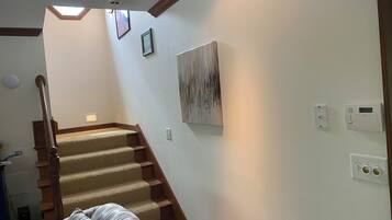 Un escalier avec des marches recouvertes de moquette et des rampes en bois, menant à un palier doté d’un luminaire au plafond et d’un thermostat fixé au mur.