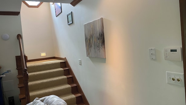 A staircase with carpeted steps and wooden handrails, leading to a landing with a ceiling light and a wall-mounted thermostat.
