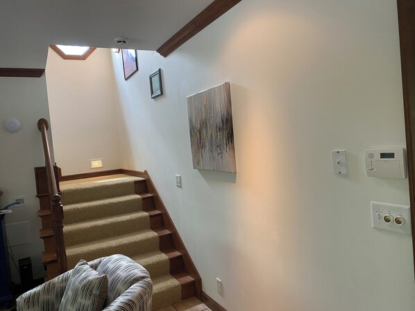 A staircase with carpeted steps and wooden handrails, leading to a landing with a ceiling light and a wall-mounted thermostat.