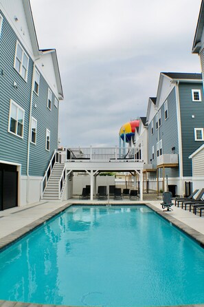 En swimmingpool med en hvid fliseterrasse, omgivet af bygninger med altaner og vinduer.