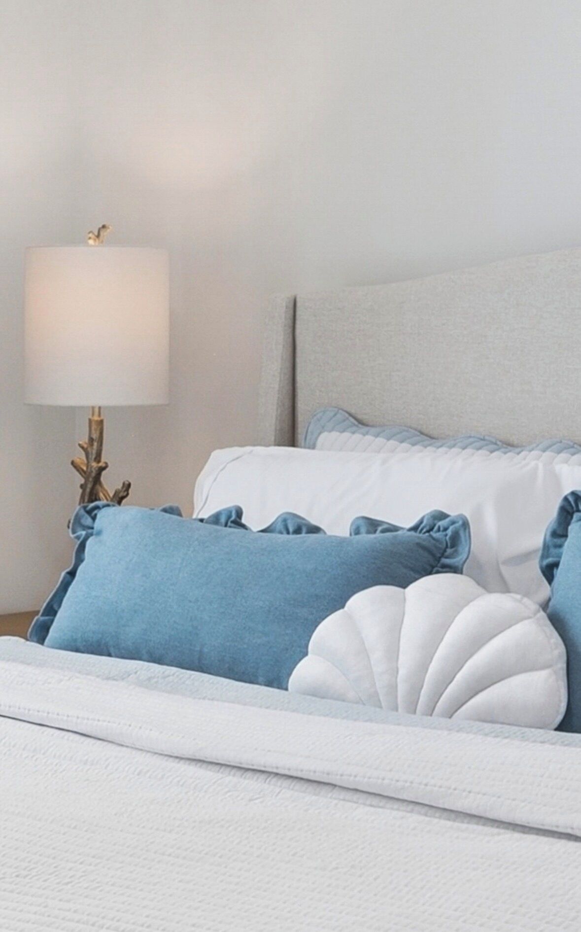 Ein Bett mit weißem Kopfteil, blau-weißen Kissen und einer Nachttischlampe mit Holzfuß.