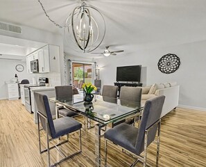 Eine moderne Küche mit weißen Schränken, einem Essbereich mit Glastisch und Metallstühlen, einem Deckenventilator und einem Flachbildfernseher.