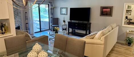 A modern living room with a glass dining table, sofas, a TV, and a ceiling fan.