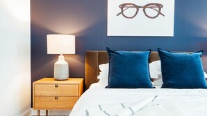 A bedroom with a bed, two pillows, a nightstand with a lamp, and a wall-mounted picture of glasses. - Landing Amazing Studio, Game Room, Gym (New Rochelle)