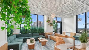 Eine moderne Lounge mit einem grünen Sofa, Holztischen, Korbstühlen und großen Fenstern mit Blick auf die Stadt.