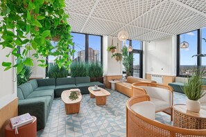 Eine moderne Lounge mit einem grünen Sofa, Holztischen, Korbstühlen und großen Fenstern mit Blick auf die Stadt.