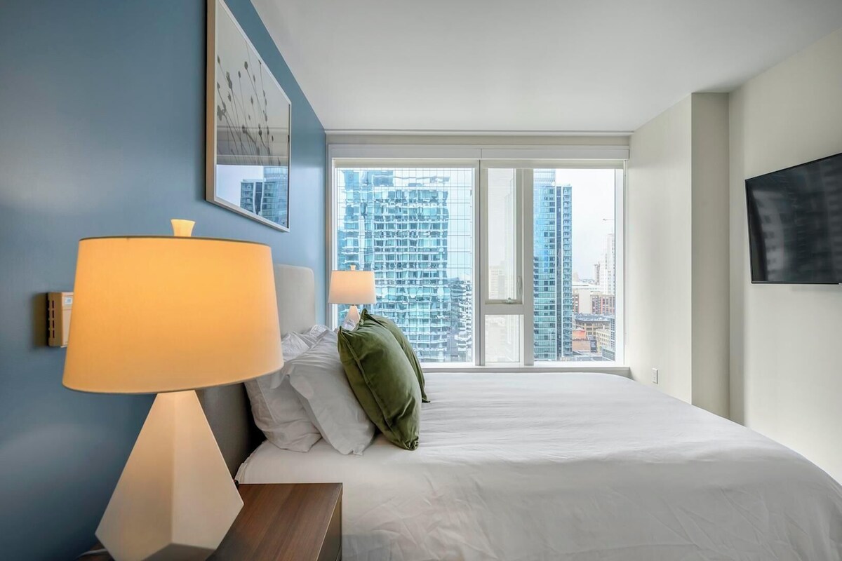 Ein Hotelzimmer mit einem großen Bett, zwei Nachttischlampen, einem grünen Kopfkissen, einem Flachbildfernseher an der Wand und einem Fenster mit Blick auf die Stadt.
