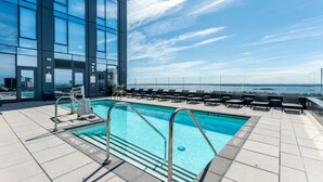 Ein Pool auf dem Dach mit klarem, blauem Wasser, umgeben von Liegestühlen und einem Glasgeländer, mit Blick auf ein Gewässer.
