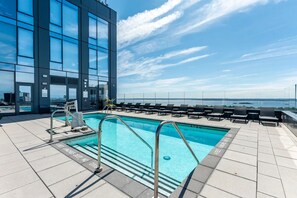 Ein Pool auf dem Dach mit klarem, blauem Wasser, umgeben von Liegestühlen und einem Glasgeländer, mit Blick auf ein Gewässer.
